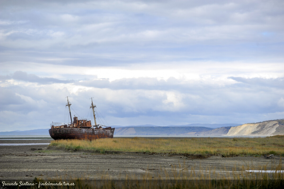 de fondo acantilados, en primer plano el lecho pantanoso del mar y en el medio un barco encallado y con oxido viejo. Rio grande , cercano a Ushuaia.